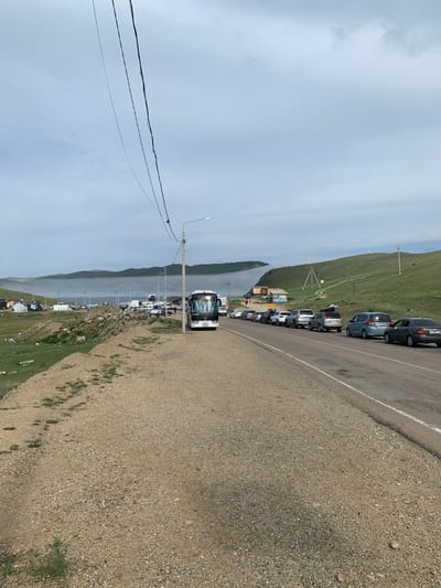 A queue of cars and a bus are seen on a road near the city of Sakhiurta, Russia, on a foggy day. The road is paved and has a white line marking the lanes. On the right side of the road, a row of cars are lined up, including a blue minivan and a dark sedan. A white bus is parked on the side of the road, facing towards the queue of cars. On the left side of the road, there are several buildings and tents, as well as a rocky terrain covered with grass. In the background, hills are visible through the fog. The sky is cloudy and overcast.
