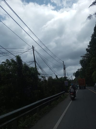 The image depicts a roadside scene in Gubbisamamidi, India, observed under an overcast sky with dense grey clouds. In the foreground, a metallic guardrail extends along the left edge of an asphalt road, which features a solid white line marking the shoulder. The road curves slightly to the right in the mid-distance. A dark-colored scooter, carrying two individuals (one in a blue helmet), travels away from the viewer on the left side of the road. Further ahead, a large orange truck with an open bed is visible in the right lane, followed by other vehicles in the distance. The left side of the road is characterized by dense green vegetation and several wooden utility poles supporting a complex, tangled network of overhead electrical or communication wires. Lush, dark green trees line the right side of the road, extending upwards to form a dense hillside backdrop.