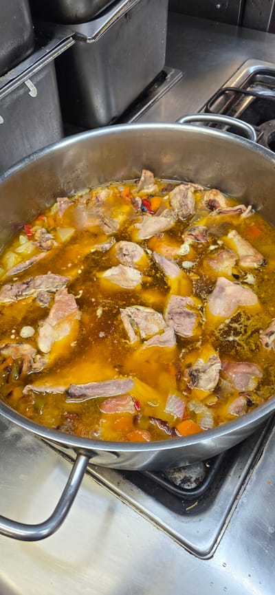 A close-up, high-angle shot shows a large pot of what appears to be a hearty stew or soup, filled with pieces of meat and vegetables, simmering on a commercial gas stovetop. The broth is a rich, golden-yellow color, with visible chunks of pale meat, diced orange carrots, and possibly white onions and red peppers. The metal pot has two sturdy handles and sits on a stainless steel surface that shows signs of use, with some minor staining and grease marks around the stovetop burners. The background is dominated by stainless steel surfaces and containers, typical of a professional kitchen environment. The scene suggests a food preparation setting, likely in a restaurant or commercial kitchen. No people are visible in the image. The location is identified as Sallent de Gállego, Spain.