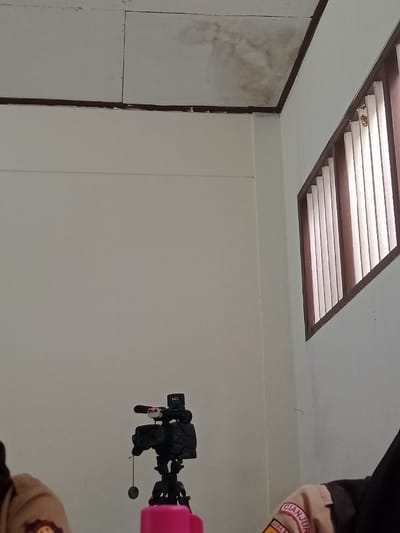 The scene depicts an interior room in Kawungluwuk, Indonesia. The room has light-colored walls and a panelled ceiling. The ceiling in the upper right quadrant exhibits a prominent, irregular dark stain, consistent with water damage, and is bordered by a dark brown trim along the wall junction. A rectangular window on the right wall features vertical brown wooden slats, allowing diffused light into the space. A black video camera, marked "HD" and equipped with a red indicator light on an accessory, is mounted on a black tripod, positioned centrally in the lower part of the frame. A small, dark, circular object hangs from the camera's left side. Partially visible in the bottom center is a pink container, likely a cup or bottle. On the far left, the shoulder and upper arm of an individual wearing a light brown uniform with a dark emblem are present. On the far right, the shoulder and upper arm of another individual are visible, wearing a similar light brown uniform with a sleeve patch displaying "CIANJUR" and other text, along with a dark head covering.