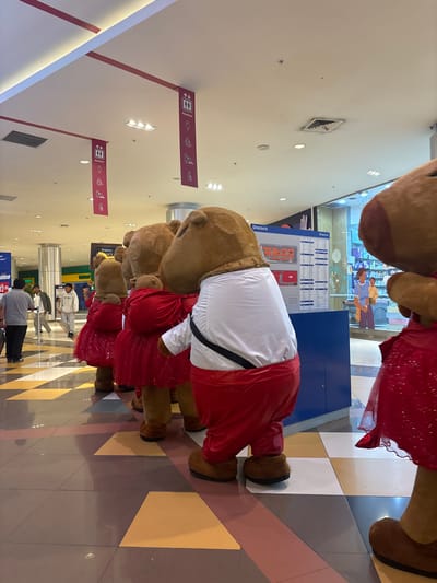 A photograph shows a row of costumed characters in what appears to be a shopping mall. The characters, presumably bears, are wearing red and white outfits. The mall's directory sign is visible behind them, along with general store information and a directory map. People are walking in the background. Overhead signs indicate the location of elevators ("Ascensor"), and the floor is tiled in a geometric pattern of brown, white, and yellow. The specific location within San Juan de Miraflores, Peru, cannot be determined solely from the image.