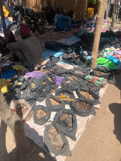 This media file captures a vibrant outdoor market scene, likely in Dull A, Nigeria. The primary subjects are people and a variety of goods displayed for sale. The setting is a dusty, sun-drenched area, suggesting a warm, possibly dry climate and a daytime time of day, indicated by the strong shadows cast by overhead objects and people.

Various objects are prominent, including a large assortment of colorful flip-flops and sandals neatly hung or displayed, suggesting a footwear vendor. Beside these, an array of black plastic bags filled with what appears to be dried goods, soil, roots, and possibly medicinal herbs are laid out on a white tarp. Some of these bags are partially open, revealing their contents. Other items include a motorcycle, suggesting transportation or goods delivery, and various pieces of clothing or textiles, some of which are brightly colored, like purple and yellow bags, and others appear to be sacks. A large piece of driftwood is positioned across the foreground.

The activity taking place is clearly a commercial transaction, with vendors displaying their wares and potential customers present, although the focus is more on the goods than the interactions. The overall impression is one of a bustling marketplace, with a sense of daily commerce. The strong sunlight and shadows imply it's a clear day. The general atmosphere seems lively and active, typical of a market. There is no visible text in the image.