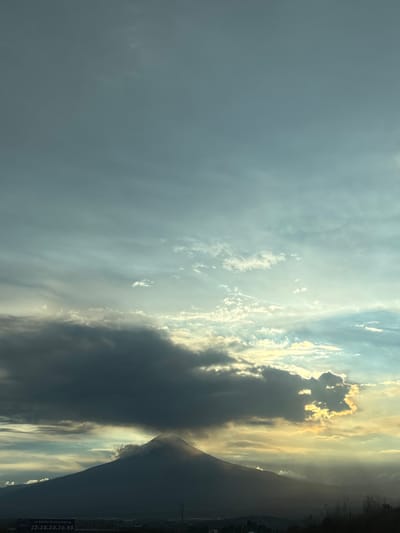 The image captures a silhouetted mountain peak, possibly a volcano, under a vast sky filled with dramatic clouds. The lower part of the image shows a dark, mountainous terrain, with the peak of the mountain prominently featured. The sky transitions from a deep blue at the top to lighter shades with wisps of clouds illuminated by what appears to be sunset or sunrise. Darker, more substantial clouds are gathered around the mountain's peak, with a bright, yellowish light breaking through in places, suggesting the sun is either setting or rising behind the clouds. The bottom edge of the image reveals a dark silhouette of a treeline and possibly some low-lying structures, hinting at a populated area at the foot of the mountain. In the very bottom left corner, a sign is visible with text in Spanish, including what appears to be a phone number and the phrase "SE RENTA ESTE ESPACIO." This suggests an urban or semi-urban environment bordering the natural landscape. The location is specified as the city of San Bernardino Chalchihuapan, Mexico.