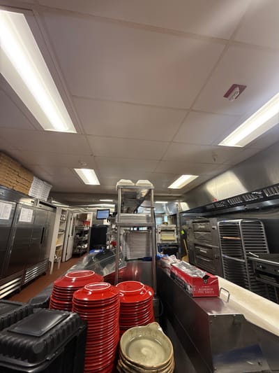 The image depicts a commercial kitchen setting, likely in a restaurant or food service establishment, in West Ocean City, Maryland, United States of America. Visible are stacks of plates (red and speckled tan) on a metal shelf, along with a box of aluminum foil sheets. There are metal shelving units with plates, and a dishwasher in the background. Further back is what seems to be more food preparation and storage areas. The general environment appears to be clean and well-lit with fluorescent lighting fixtures.