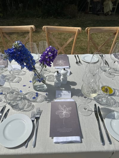 The image depicts a set table outdoors, likely at a wedding or formal event. The tablecloth is light grey, and the place settings include white plates, silver cutlery, multiple wine glasses, and menus with a floral design. Each setting also has a small name card and a salt and pepper shaker. A glass vase with blue and purple flowers serves as a centerpiece. The chairs are light wood with a cross-back design. The location appears to be outdoors on a grassy area. It's impossible to confirm the location as Cambridge from the image alone.