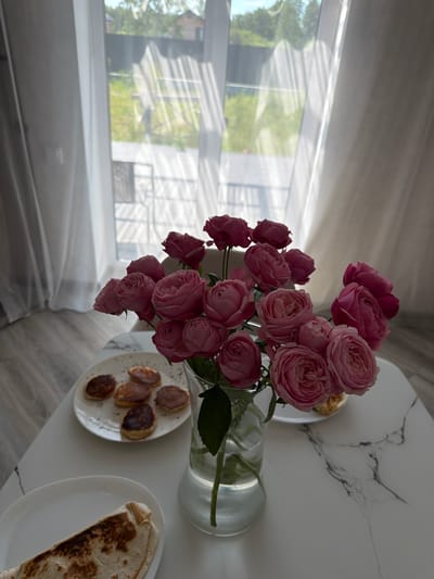 A still life photograph features a bouquet of pink roses in a clear glass vase on a white marble-patterned table. To the left of the vase, a white plate holds several small, round pancakes. Another white plate below it contains a folded flatbread wrap. In the background, sheer white curtains are drawn, revealing a blurred view of greenery and a fence outside, suggesting a residential area in Malinovka, Russia. The lighting indicates it is daytime. There are no people visible in the image.