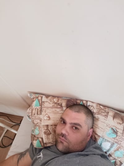 A man is lying in bed, looking at the camera. He has a shaved head and a short beard. He is wearing a gray hoodie. The pillow he is lying on has a pattern of hearts and other shapes. The room appears to be in a residential setting. There is a white wall and what appears to be a bed frame and some electrical cords in the background. The image does not contain any information about the city of Babylon, Czechia.