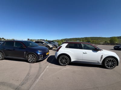 The image displays an outdoor parking lot under a clear, bright blue sky on a sunny day. Two modern compact crossover-style cars are parked prominently on the asphalt in the foreground.
On the left, a dark blue car features distinctive squared "C"-shaped LED daytime running lights at the front. It is equipped with multi-spoke wheels in a silver and black design and bears a subtle "5" emblem on its side fender. The car's front license plate, "KB 8022," is clearly visible, indicating Belgian registration.
To its right, a white car exhibits a sporty design. It has a contrasting black roofline highlighted by a red trim that runs along the side windows and roof edge. This vehicle also features pronounced black fender flares, multi-spoke black and silver wheels, and a patterned, vertical vent-like element on its front fender.
In the mid-ground and background, several other vehicles are parked. Beyond the parking area, a landscape of lush, tree-covered hills extends under the expansive blue sky. Distant infrastructure, including streetlights and a road segment, can be seen on the hillsides. No people are visible in the scene. The overall impression is one of a bright, pleasant day in a parking facility, located in Aubange, Belgium. A partially visible license plate "DW 837-TG" can be seen on a dark car in the far background on the right.