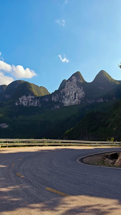 The image shows a curved section of asphalt road in Đồng Văn Commune, Vietnam. The road has yellow lane markings and a white guardrail. Mountains with visible rock formations and green vegetation are in the background. The sky is blue with scattered clouds. Shadows are visible on the road surface.