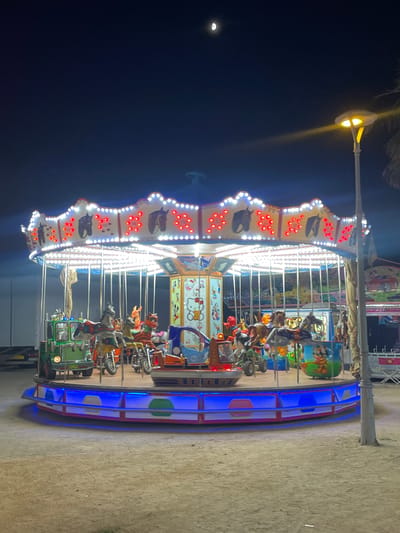 This media file shows a brightly lit carousel at night, likely at an amusement park or fair in Canet de Mar, Spain. The carousel is adorned with numerous white lights, accentuating its circular structure, and punctuated by red lights. Various animals, such as horses, and cartoon characters, including Hello Kitty, serve as the ride's seats. These seats are arranged in tiers on poles, suggesting they can move up and down and around. Some of the seats appear to be designed as vehicles, like a small green truck.
The scene is set against a dark night sky, with a sliver of the moon visible in the upper left corner. A streetlamp with a warm yellow light illuminates a portion of the sky and a palm frond, adding to the evening ambiance. The ground is a sandy or gravelly surface, typical of outdoor recreational areas. The carousel itself has a colorful base with blue lights, indicating it's an active amusement ride.
While no people are clearly depicted in detail, the presence of the carousel implies that people are likely using or waiting to use the ride, creating a festive and entertaining atmosphere. The overall mood is cheerful and lively, characteristic of a nighttime fair or carnival. The image captures the dynamic and vibrant nature of such an attraction.