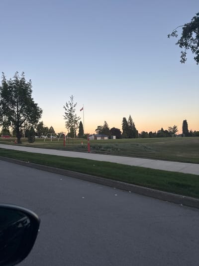 The image depicts a park scene at dusk, with a clear sky transitioning from a soft blue to a warm orange hue near the horizon, suggesting the sun is setting. A paved road runs along the right side of the frame, bordering a grassy area and a sidewalk. Several trees are scattered throughout the park, with one large, mature tree prominently featured in the foreground. In the mid-ground, a Canadian flag waves on a flagpole. Orange traffic cones are placed along the sidewalk, possibly indicating a temporary closure or event. In the distance, there are glimpses of buildings and a soccer goal, further defining the park's setting. A portion of a car's side mirror is visible in the lower right corner of the image. The overall mood is peaceful and serene, characteristic of a quiet evening in a suburban park. There is no visible text in the image.