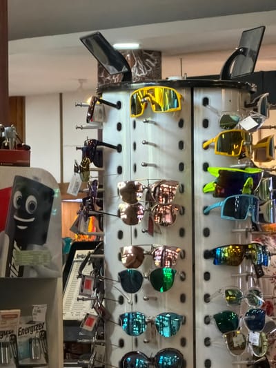 The image captures an interior retail scene within a shop in Benoa, Indonesia. A prominent, multi-sided, light-colored rotating display rack for sunglasses occupies the central and right portions of the frame. This rack showcases numerous pairs of sunglasses, characterized by diverse frame colors (including yellow, black, neon green, and light blue) and various mirrored lens finishes (yellow, green, blue, rose gold, and dark tints). Styles range from large, wrap-around designs to more conventional shapes. Many sunglasses have attached tags, likely indicating price or brand. The top of the display rack features two dark, reflective, angled components.

In the left foreground, a white display structure presents a black, rectangular object featuring a cartoon-like face with large white eyes and a wide smile, accessorized with what appears to be a white shirt and a striped tie. Directly below this, a lower shelf displays blister packs of "Energizer" brand batteries. The background reveals additional retail shelving and a patterned wall segment. Overhead, fluorescent lighting illuminates the space, and a white ceiling fan is partially visible in the upper left. No individuals are present in the scene.