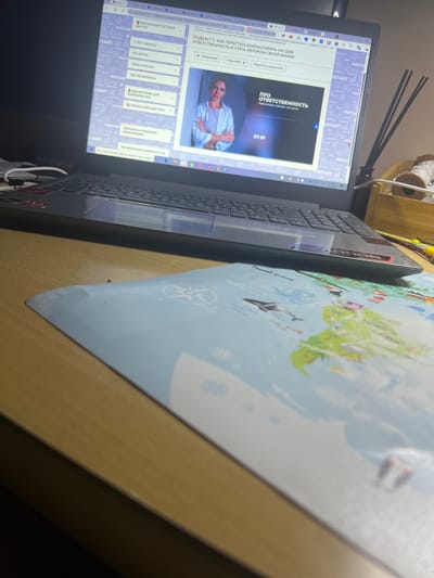A laptop is open on a wooden desk, displaying a video course. The course title "ПОДКАСТ 2. КАК ПЕРЕСТАТЬ БОЯТЬСЯ БРАТЬ НА СЕБЯ ОТВЕТСТВЕННОСТЬ И СТАТЬ АВТОРОМ СВОЕЙ ЖИЗНИ" (Podcast 2. How to Stop Being Afraid to Take Responsibility and Become the Author of Your Own Life) is visible, along with a woman speaking in the video. The left side of the screen shows a navigation menu with options like "БЕСПЛАТНЫЙ ГОСТЕВОЙ ДОСТУП" (Free Guest Access) and "ВСЕ КУРСЫ" (All Courses). To the right of the laptop, a wooden box with aroma sticks and a spool of white thread are present. A light blue mat with illustrations of the world map and sea creatures is partially visible in the foreground. The overall environment suggests a home office or study area. The image does not contain any specific information about the location being in Водник, Russia.