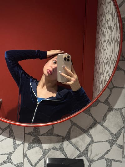A woman is reflected in a round, red-framed mirror. She is holding a phone in her right hand, presumably taking a selfie. She is wearing a navy blue, long-sleeved zip-up shirt. The background of the reflection includes a red door and a ceiling with a geometric tile pattern. The visible background outside of the mirror shows a wall with a similar geometric tile pattern and a dark object attached to the wall. There is no visible information to verify the location as Borok, Russia.