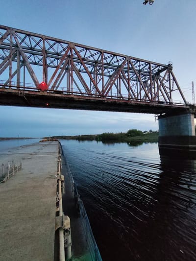 The image depicts a steel truss bridge spanning a river, likely in Tarkanovo, Russia. The bridge has a reddish-brown, weathered appearance with a complex lattice structure. A concrete pier supports one end of the bridge on the right side. Underneath the bridge, on the left, there's a concrete dock or platform extending into the river. The water appears dark and slightly choppy, with visible ripples and reflections. The opposite riverbank is visible in the distance, featuring trees and some houses. The sky is overcast, with a gradient of light blue to gray, indicating either dawn or dusk. A drone is visible near the bridge in the sky. Red lights are attached to the bridge structure.