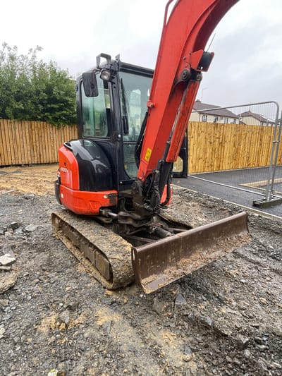 The image shows an orange and black excavator parked on a gravelly construction site. The excavator is the primary subject, with its boom and bucket visible. The background includes a wooden fence, a metal safety barrier, and some buildings that appear to be under construction. The sky is overcast, suggesting cloudy weather.

There are no people visible in the image. The scene is outdoors, likely in a construction or renovation area. The ground is covered in loose stones and dirt, with some muddy patches. The excavator appears to be stationary, with no activity taking place.

Notable details include the model number "U00374" visible on the side of the excavator. The weather appears to be overcast, and the overall impression is one of a working construction site. The location context provided is Banbridge, United Kingdom.