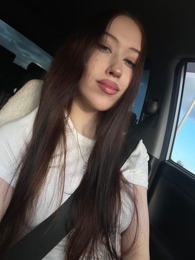 This is a selfie taken inside a car. The subject is a young woman with long, dark brown hair. She is wearing a white t-shirt and a silver necklace with a small charm. Her face is angled towards the camera, with her eyes partially closed and her lips slightly parted. She appears relaxed and is leaning back against the car's headrest, which has a fuzzy, cream-colored trim. A seatbelt is visible across her chest. The background shows the interior of the car, including the window and a glimpse of the sky and trees outside, suggesting it is daytime with clear weather. The lighting suggests it might be late afternoon or early evening due to the warm tones on her skin. There is no visible text in the image.