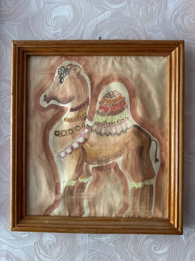 A framed watercolor painting is centered in the image against a textured white background. The painting depicts an abstract brown figure with ornate decorations. The figure has a decorated headpiece with red, orange, yellow, and green details. Markings and patterns are visible on the body. The painting is contained within a wooden frame with a gold finish. The scene is located in the city of Sayansk, Russia.