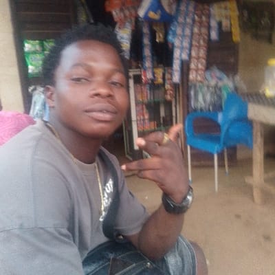 A medium close-up portrait captures a young Black man outdoors, likely in Otuocha, Nigeria. He is positioned slightly to the left, looking directly at the camera with a neutral to slightly smiling expression. He has short, dark, curly hair and is wearing a grey t-shirt with a gold-colored chain around his neck, a dark wristband or watch on his left wrist, and dark shorts or trousers. His right hand is raised, making a gesture with his index and pinky fingers extended, while his thumb is also extended, possibly indicating a "rock on" sign.
The background reveals a retail environment, characteristic of a small shop or market stall, under what appears to be a roof or awning. Numerous colorful, packaged goods, likely snacks or candies, hang from shelves or strings, though they are out of focus. A distinctive blue plastic chair is visible on the right side of the frame, alongside part of a wooden table or counter on which a plastic bottle sits. The natural, bright lighting suggests it is daytime. The scene conveys a casual moment in a local commercial setting. No specific text is clearly legible in the image.