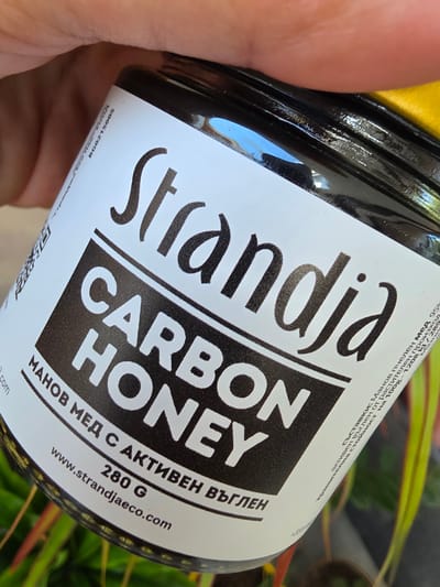 The image shows a close-up of a jar of honey with a label. The label features the brand name "Strandia" in a stylized font, followed by "CARBON HONEY" in bold, white, block letters on a black background. Below this, in Bulgarian, it reads "МАНОВ МЕД С АКТИВЕН ВЪГЛЕН" (Manov honey with active carbon). Further down, it states "280 G" and a website address "www.strandjaeco.com". A QR code is visible on the left side of the label, and some text in Bulgarian, including ingredient information and a production batch number "BG0215005", is present on the right side of the label, partially obscured. The jar is held by a hand, with a blurred background of green and yellow foliage. There is no discernible information or context to place this image specifically in the city of Indzhe Voyvoda, Bulgaria.