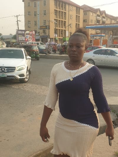 A woman stands outdoors in front of a street scene. She wears a two-toned navy and white dress and holds keys in one hand and a phone in the other. Behind her is a building with multiple windows and shops at street level, visible through traffic including cars and motorcycles. A billboard is visible, advertising a restaurant. The sky is overcast.