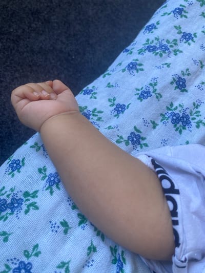 The image shows a close-up of a baby's arm resting on a light blue fabric with a floral pattern of blue flowers and green leaves. The baby's hand is visible at the top left of the frame, with fingernails clearly defined. The baby's arm is smooth and lightly tanned. A white garment with black lettering, possibly a brand logo, is visible on the right side of the arm. The background appears to be a dark, textured surface, possibly upholstery or fabric. The scene is likely indoors or in a shaded outdoor area, given the soft lighting. The image provides no information that would allow for its localization in Arbagish, Uzbekistan.