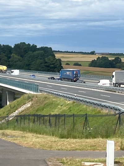 The image captures a section of a multi-lane highway under an overcast sky. In the foreground, a grassy embankment is visible with a black safety fence. The highway itself features several vehicles, including a blue truck with "BET" branding on its side, a white van, and a white semi-truck. A yellow tanker truck is partially visible on the far left of the highway. The highway is elevated by concrete supports, suggesting it is a viaduct or overpass. In the background, rolling hills with patches of trees and agricultural fields are discernible, indicative of a rural or semi-rural setting. The location is identified as Momberg, Germany, suggesting this highway is part of the German road network. There are no people clearly visible in the image. The overall scene depicts a typical highway environment with various commercial vehicles.