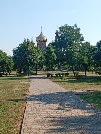 The image shows a paved walkway in a park on a sunny day. The walkway is made of light-colored bricks and curves to the right, leading the eye towards the background. On either side of the walkway, there is a grassy area with scattered trees and bushes. The trees are lush and green, casting shadows on the path. In the background, a church with golden domes is visible through the trees. The sky is clear and blue, indicating fair weather. The overall scene is peaceful and serene, suggesting a quiet day in a park. There are a few people visible in the distance, walking in the park, and some birds are scattered on the grass. The time of day appears to be morning or afternoon, given the angle of the shadows. The presence of the church with golden domes suggests the location might be a religious or historically significant site within Pokrov, Ukraine. No visible text is present in the image.