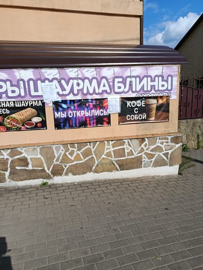 The image shows the exterior of a food establishment, likely a cafe or fast-food restaurant, located on the corner of a building in Смілавіцкі сельскі Савет, Belarus. The building has a beige facade and a dark brown corrugated metal awning. A large banner with purple lettering across the top reads "ПЫШНЫЕ БЛИНЫ" (fluffy pancakes). Below this, there are three posters. The leftmost poster features an image of a shawarma wrap and text in Russian. The middle poster has the text "МЫ ОТКРЫЛИСЬ!" (We are open!) in large white letters against a colorful, abstract background. The rightmost poster displays a cup of coffee with the text "КОФЕ С СОБОЙ" (coffee to go) in white letters on a dark background. The building's lower section is decorated with a stone pattern. A brick-paved walkway runs in front of the building. The sky above is blue with scattered white clouds. A decorative iron fence is visible on the right side of the image.