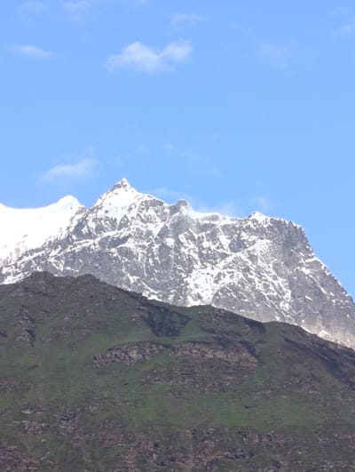 An extreme low-angle medium shot captures a vast, snow-capped mountain range beneath a bright blue sky dotted with wispy clouds. The lower portion of the image reveals a foreground of dark, vegetated slopes. The mountains ascend dramatically, with their peaks heavily dusted with snow. The detailed texture of the rocky terrain and the patches of green vegetation on the lower slopes are clearly visible. The scene is devoid of any human presence or man-made structures, emphasizing the natural grandeur of the landscape.