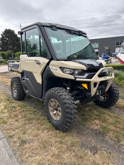 The image shows a tan Can-Am Defender UTV parked on a grassy area next to a paved surface, likely a road or driveway. The UTV is oriented sideways in the frame. The vehicle has large, knobby tires and a protective front bumper. The windshield is wet, indicating recent rain.
In the background, a modern, dark-colored building with large windows and a "SHIPPING & RECEIVING" sign is visible. The sky is overcast and appears to be a grey, cloudy day. A few power lines are also visible against the sky. The ground is covered in dry, brownish-yellow grass with some patches of green, suggesting a potentially dry climate or season. There is also some dirt and debris on the ground.
No people are visible in the image. The overall mood is neutral, with no strong emotional cues. The time of day is not clearly indicated but the overcast sky suggests it could be anytime during daylight hours. The presence of the building and power lines suggests an urban or semi-urban setting within the Township of Langley, Canada. Visible text includes "DEFENDER", "CAN-AM", and "SHIPPING & RECEIVING".