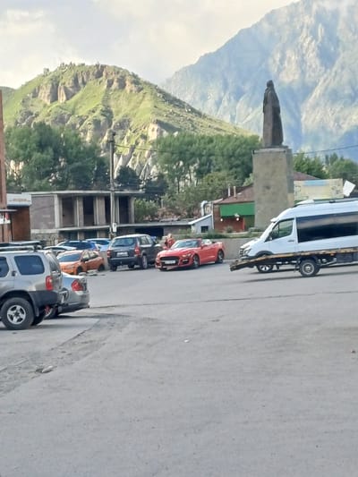 In Stepantsminda, Georgia, a diverse collection of vehicles occupies a paved area, with a prominent statue of a cloaked figure on a pedestal situated in the mid-ground, overlooking the scene. A red convertible sports car is parked to the right of a dark SUV, with an orange compact car positioned behind it. A silver SUV is visible in the foreground to the left. A white tow truck, carrying a flatbed trailer, is positioned to the right of the red convertible. In the background, a partially constructed building stands, and beyond that, lush green trees line the base of a rugged, mountainous landscape. The sky is overcast, suggesting a diffused light.