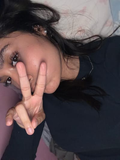 This is a close-up, rotated image of a person lying down, possibly in bed. The subject has dark hair that is spread around their face and shoulders. They are wearing a black long-sleeved shirt. Their face is partially obscured by their hand, which is held up in a peace sign gesture in front of their eyes. The person appears to be looking directly at the camera.
The setting seems to be indoors, given the presence of what looks like bedding with a light-colored pattern, and the overall lighting suggests it could be nighttime or a dimly lit room. There is no visible text in the image. The mood appears casual and perhaps playful due to the peace sign gesture. The person's eyes are visible, and they are dark and appear to have mascara on their lashes. A delicate silver necklace with a small charm is visible around their neck. The overall impression is an intimate and personal selfie.