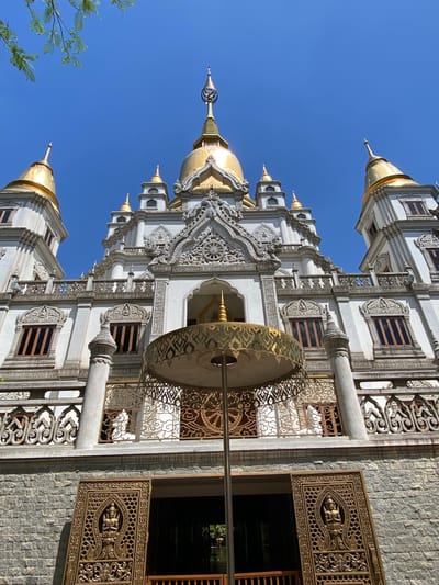 The image shows the Kim Cang Tu Buddhist temple in Dĩ An, Vietnam, taken from a low angle looking upwards. The temple features white walls and multiple golden domes. The building has ornate carvings and decorations. A large golden parasol is visible in the foreground, partially obscuring the entrance. The sky is clear blue.