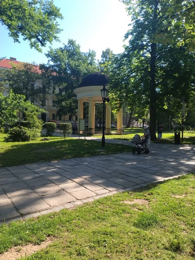 The image captures a sunny day in a park, likely in Písek, Czechia, given the kiosk's inscription. In the foreground, a paved walkway with stone tiles is bordered by a strip of green grass. A person wearing a light-colored hat and striped shirt is pushing a gray baby stroller along the walkway. Behind them, a mature tree casts dappled shadows on the path. Further back, a classical-style yellow rotunda with a dark dome stands prominently. The rotunda has several columns and a sign with text, which appears to read "SIEM CO HOR." Trees with lush green foliage surround the rotunda and the park. In the background, a multi-story building with a red-tiled roof and cream-colored facade is visible, suggesting an urban park setting. Another person stands near a trash can under the shade of a tree. The overall scene depicts a peaceful, everyday moment in a public space.