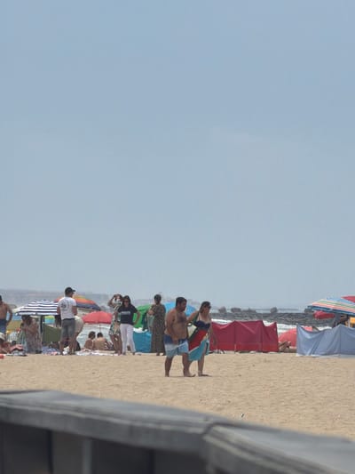 The image depicts a crowded beach scene on a sunny day, likely in Espinho, Portugal, given the context. The foreground is dominated by a wooden railing, obscuring the lower portion of the beach. The sandy beach is populated with numerous individuals, some in swimwear, others in casual clothing. Various beach umbrellas, including striped and solid colored ones, are scattered across the sand, providing shade. Several beach shelters, some red and some blue, are set up, with people visible inside or near them. In the mid-ground, a couple walks hand-in-hand, the man wearing blue shorts and the woman in a blue bikini top and a colorful sarong. Other people are seen standing, sitting, and interacting amongst themselves. The background shows the ocean with waves breaking against rocky outcrops. The sky is a pale, hazy blue.