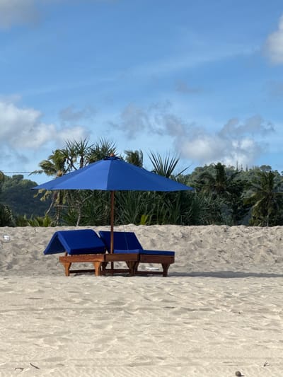 A beach scene is captured with a blue umbrella casting a shadow over two blue-cushioned beach chairs on a white sandy shore. Tropical foliage, including palm trees, is behind the chairs. The sky is blue with scattered clouds. The scene is likely in Selong Belanak, Indonesia.