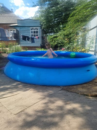 A child with blonde hair is playing in a bright blue inflatable swimming pool filled with water. The child is facing away from the camera, with their right arm extended. A green ball floats in the pool to the right of the child. The pool is situated outdoors on a sandy area, with some dirt and fallen leaves visible on the ground around it. In the background, there is a small wooden structure painted blue with a white-paned window and clothes hanging on a line. A grey-tiled roof of a building is visible behind the wooden structure, and dense green foliage forms a backdrop on the right side of the image, with a corrugated metal fence partially visible. The scene suggests a backyard or a communal outdoor space in a residential area, possibly in Romny, Ukraine, given the typical architecture and vegetation. The sky above is partly cloudy.