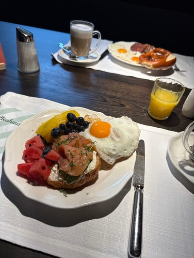 A breakfast scene is captured on a dark wooden table, covered partly by a white textured placemat. In the foreground, a white ceramic plate holds a variety of breakfast items: sliced watermelon, blueberries, a piece of what appears to be mango, a croissant topped with cream cheese, smoked salmon, and fresh herbs, and a fried egg with a runny yolk. A silver knife rests to the right of the plate. Further back on the table, another white plate contains two fried eggs, some bacon, and a pretzel. A glass of orange juice sits in the mid-ground. In the background, a cup of coffee with foam sits on a saucer, next to a glass sugar dispenser. The setting appears to be a restaurant in Mayrhofen, Austria, suggested by the "Neuhaus" branding visible on the placemat. The lighting is dim, with a strong light source illuminating the breakfast plate.