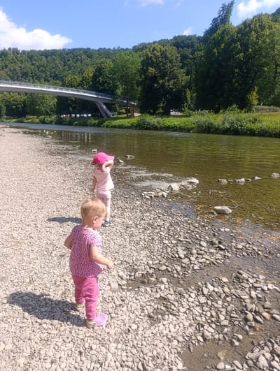 Two young children are standing on a pebble-strewn riverbank. The child in the foreground, facing away from the camera, wears a pink t-shirt with dark polka dots and pink pants, along with bright pink shoes. The second child, slightly further away and also facing away, wears a pink outfit and a pink hat. They are near the edge of a calm, greenish river. In the background, a modern, white pedestrian bridge spans the river, connecting to a heavily wooded hillside. The sky above is blue with scattered white clouds. This scene is located in Teplice nad Bečvou, Czechia.