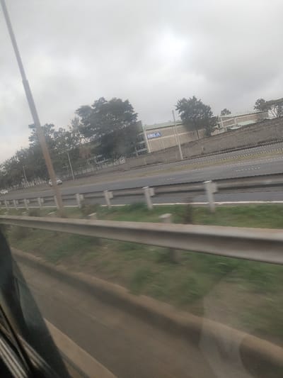 The image presents a view from a moving vehicle along a multi-lane highway in Gatongora ward, Kenya, under an overcast sky. In the immediate foreground, a grey metal guardrail runs horizontally across the lower portion of the frame, with blurred green grass beneath it, indicating motion. Beyond this, a wide asphalt highway extends into the distance, featuring at least three lanes in the direction of the viewer and at least three opposing lanes separated by a grassy median. White lane markings are discernible on the asphalt. Within the median, intermittent white posts and additional shorter guardrails are visible. Two indistinct white vehicles are present in the distance on the opposing carriageway. Further beyond the highway, a tall, grey concrete or stone wall runs parallel to the road. Behind this wall, several large industrial or commercial buildings with light-colored walls and roofs are situated. One prominent building displays a large blue sign with white lettering that reads "CMAX 4." Numerous deciduous trees line the highway and are visible behind the buildings. Several vertical utility poles or lamp posts are distributed along the roadside. No individuals are depicted in the scene.