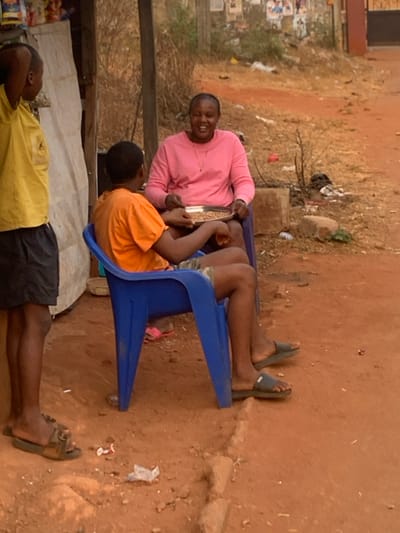 The image captures an outdoor scene, appearing to be a casual gathering or roadside activity. The ground is reddish-brown dirt or sand, with sparse dry vegetation and some litter scattered in the background.
In the center of the frame, two individuals are seated on blue plastic chairs. One person, wearing a pink long-sleeved shirt, sits facing towards the left of the image (as if looking slightly towards the camera's original position). This person is smiling broadly and holding a metallic tray or bowl filled with light-colored grains or seeds. Across from them, facing the person in pink, is another individual wearing an orange t-shirt. This person's arms are positioned around the tray, suggesting they are also engaging with its contents, possibly sifting, sorting, or sharing the grains.
To the right of the person in pink, a pair of legs stretched out on a blue plastic chair is visible, wearing dark flip-flops, indicating another person is seated there, largely out of frame. In the foreground, partially obscured on the left, a third person is standing with their back mostly to the viewer. This individual is wearing a yellow t-shirt and dark shorts, with their head slightly tilted back, as if looking up or resting. A white or light-colored tarp or sheet is visible behind this person, possibly part of a makeshift shelter or stall.
The lighting suggests it is daytime, possibly late afternoon or overcast, as there are no harsh shadows. The overall mood appears relaxed and social, with the prominent smile of the person in pink conveying a sense of contentment or friendly interaction. No clear text is visible in the image.
