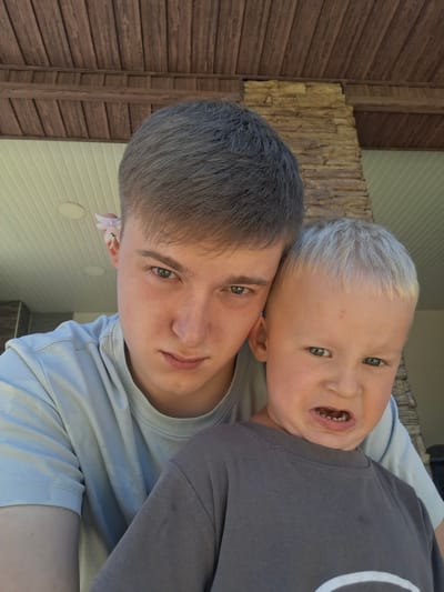Two individuals are positioned closely together in a selfie-style photograph. The person in the foreground, on the right, is a young child with light-colored hair, wearing a dark gray t-shirt. They are making a grimacing expression with their mouth open, revealing their teeth. To the left and slightly behind the child is an older individual with dark hair, wearing a light blue t-shirt. A small pink flower is tucked behind their left ear. The background consists of a wooden ceiling structure and a stone column, suggesting an outdoor or semi-outdoor setting. The image does not provide specific visual cues to pinpoint its location within the city of Kara-Balta, Kyrgyzstan.