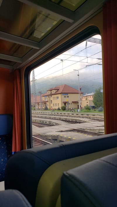 The image depicts a view from inside a train carriage, looking out at the Margecany train station in Slovakia. The foreground shows the interior of the train with dark blue seats and yellow accents. Beyond the window, the train station is visible, featuring a light yellow building with a red tiled roof. The building has multiple windows and signage indicating "MARGECANY". Train tracks and platforms are prominent in the mid-ground. Overhead, electrical wires and supports for the train lines are visible against a cloudy sky. In the background, a forested hillside rises under a hazy sky. Two individuals are visible on the station platform. One appears to be standing near a bench, and another is further away, possibly walking. The overall scene captures a moment at a rural Slovakian train station.