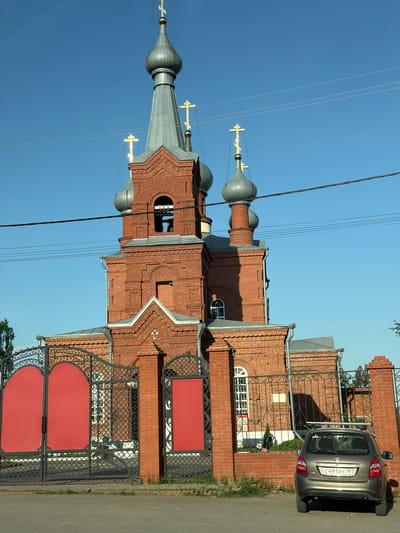 The image shows a red brick Orthodox church with multiple gray domes topped with golden crosses, juxtaposed against a clear blue sky. Power lines are visible, intersecting the sky. To the right of the church, there's a brick wall with an ornate black metal fence featuring red panels. A silver car is parked partially in front of the wall. The location is likely in the city of Malaya Purga, Russia.