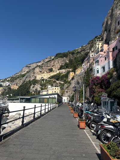 The image depicts a scenic view of the Amalfi Coast in Pogerola, Italy. A wide, paved walkway runs parallel to the rocky coastline, bordered by a black metal fence. On the left side of the walkway, large grey rocks are visible, leading to the sea. To the right of the walkway, a row of parked scooters and motorcycles are lined up along the curb. Beyond the scooters, a building with flower boxes and awnings is present, and further up the steep cliff face, colorful houses are built into the rock, characteristic of the Amalfi Coast architecture. The sky is clear and blue, indicating a sunny day. The overall scene conveys a sense of a picturesque coastal town.