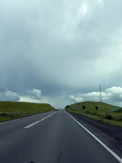 An asphalt road with double white lines runs through a landscape of rolling green hills under a cloudy sky. The road curves to the right, with a grassy embankment on the left and a hill topped with a communications tower on the right. A few cars are visible on the road in the distance. The sky is overcast with dark clouds, suggesting potential rain. The scene appears to be located in a rural or semi-rural area, possibly outside of a city like Verkhneozernoye, Russia. The overall impression is one of travel through an open, natural environment.