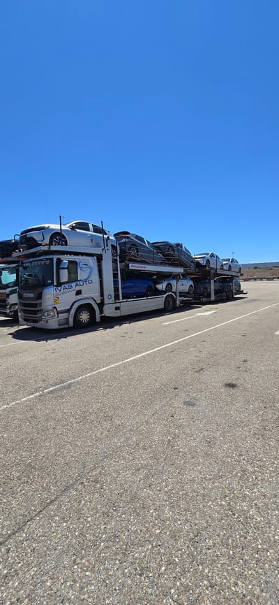 This image shows a car carrier truck filled with cars, parked on an asphalt surface next to a white line. The truck is oriented vertically in the frame, suggesting it might have been rotated for capture or display. The sky is a bright, clear blue, indicating daytime and good weather. In the background, a very distant, flat landscape is visible.

The car carrier truck is a multi-level vehicle designed to transport multiple automobiles. Several cars are clearly visible on its decks. The truck itself is predominantly white and silver, with branding that includes the text "IVAS AUTO" and a phone number. The word "SCANIA" is also visible on the front of the truck cab. One of the cars on the carrier has the text "Kässbohrer" on its side, which is a manufacturer of car transporters.

The scene appears to be a parking or staging area, possibly at a transport hub or dealership, given the presence of a car carrier. The specific location is identified as Pedrola, Spain. The bright sunlight and clear sky suggest it is a warm, sunny day. No people are visible in the image, and the focus is entirely on the truck and its cargo. The overall impression is one of efficient logistics and transportation of vehicles.
