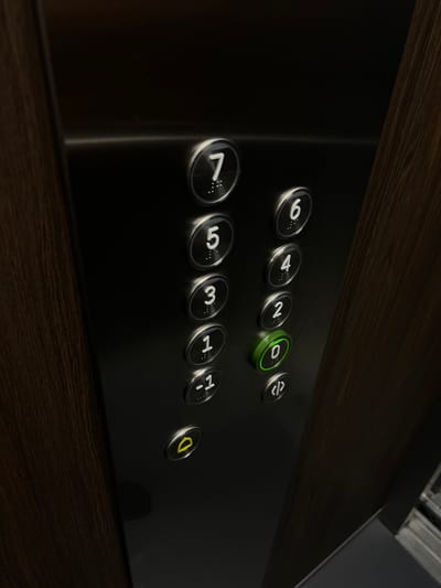 The image shows a close-up of an elevator panel. The panel is made of brushed metal and has a dark wood grain texture on the right side. There are several buttons on the panel, arranged in two columns. The left column has buttons labeled "7", "5", "3", "1", "-1", and a yellow alarm button. The right column has buttons labeled "6", "4", "2", "0", and a button with a "</>" symbol. The "0" button is illuminated green, indicating the elevator is currently on the ground floor. The lighting is dim, creating reflections on the metal surface. There are no people visible in the image. The context of Florești, Romania, is not visually represented in this image.