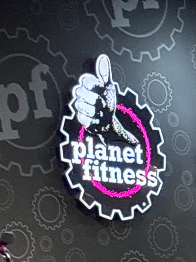 The image shows a close-up of a Planet Fitness logo. The logo features a stylized thumbs-up hand with a pink ribbon behind it, all enclosed within a gear-shaped outline. The words "planet fitness" are prominently displayed in white, stylized lettering below the hand. The background is dark gray with a subtle pattern of lighter gray gears. The overall impression is a vibrant and modern gym branding. This image was taken in Columbus, United States of America, specifically at the Planet Fitness located at 3260 Kingsmill Parkway, Columbus, OH 43229.